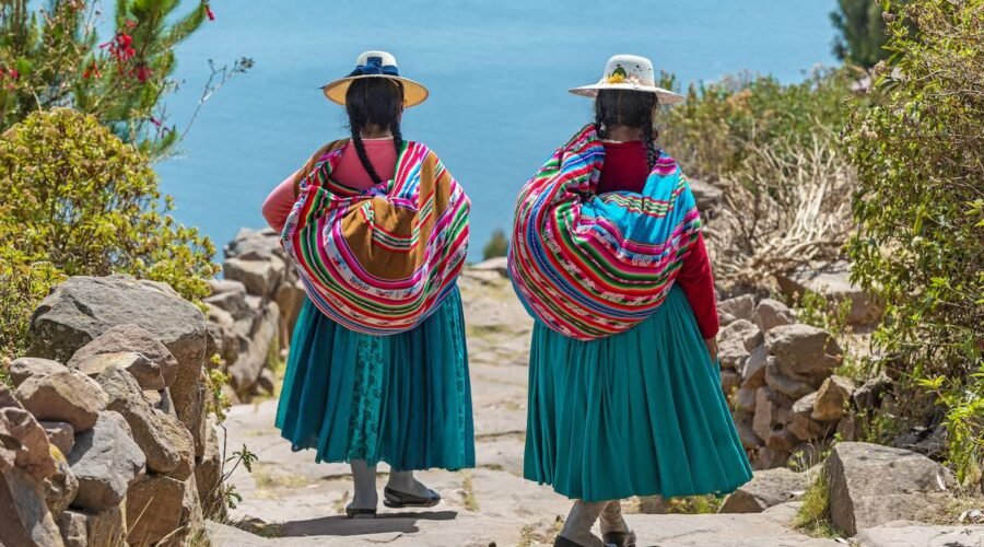 Two indigenous Quechua women in traditional clothes walking down the path to the harbor of Isla Taquile (Taquile Island) with the Titicaca Lake in the background, Peru.